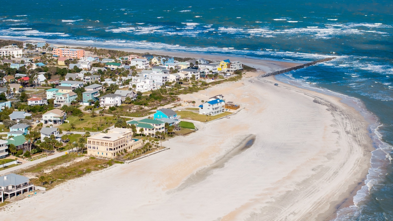 The image shows an aerial view of Vilano Beach in St. Augustine, Florida. The beach is bordered by the Atlantic Ocean, with clear distinctions between the sandy shore and the ocean waters. Buildings and structures are visible along the coastline.