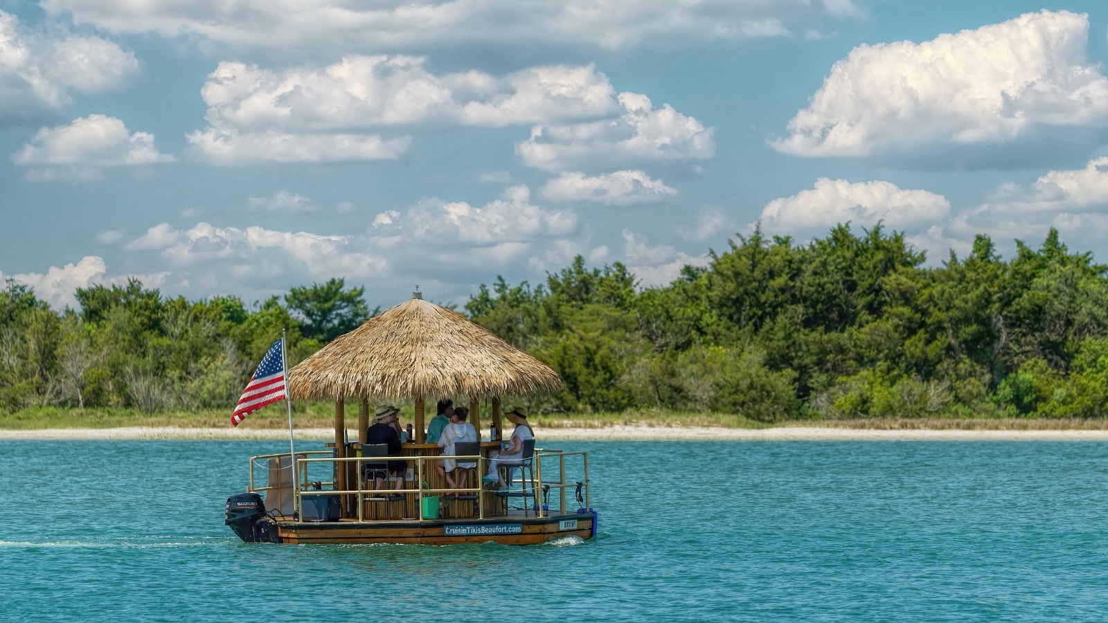 The image features a Tiki Boat Cruise in St. Augustine, showing a boat with a roof made of yellow hay floating next to the USA flag. The scene captures a leisurely boating experience with passengers on board enjoying the outing.