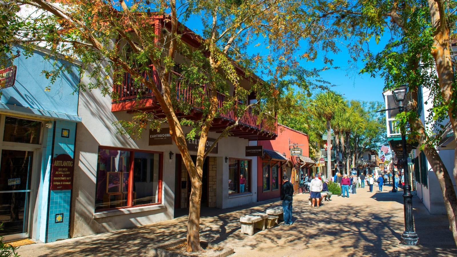 The image depicts a view of Meader Down St. George Street, showcasing people casually walking along the sidewalk. The scene includes benches placed strategically for resting, with trees providing a light shade over the area. Various shops and their signs are visible along the street.