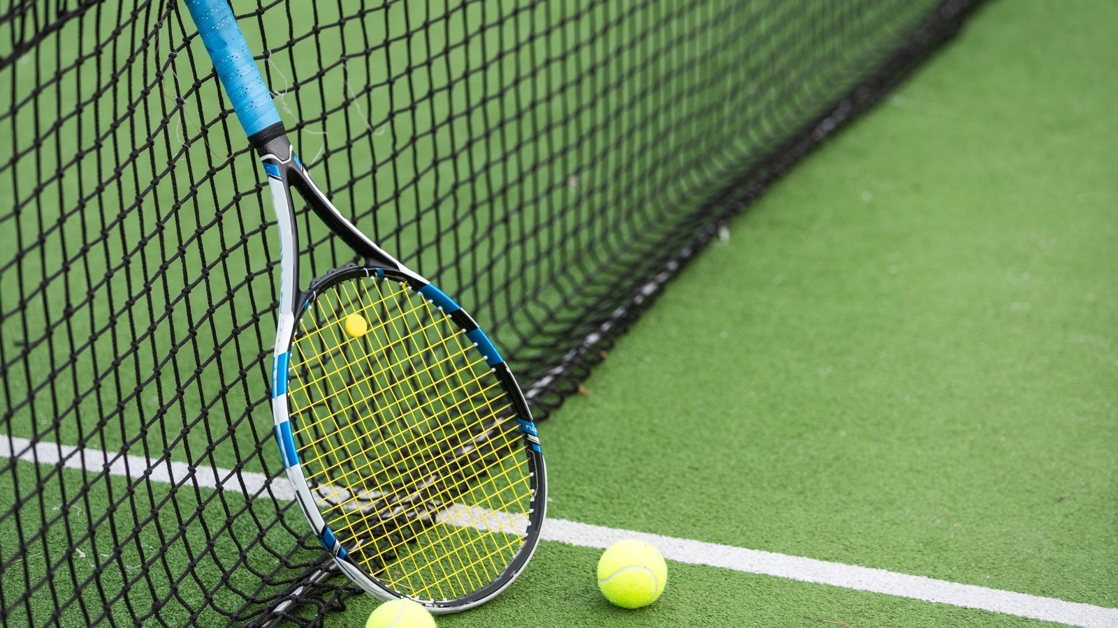 A close-up photograph depicting a tennis racket and two tennis balls on a tennis court.
