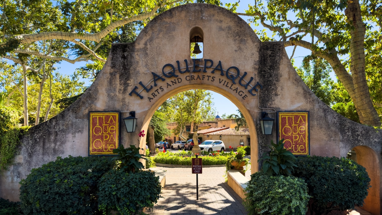 A photograph depicting the concrete entrance arch at Tlaquepaque Arts & Crafts Village. The arch prominently features the inscription 'Tlaquepaque Arts & Crafts Village'. Bushes flank the sides of the arch, while trees tower above it, and abundant vegetation surrounds the area.