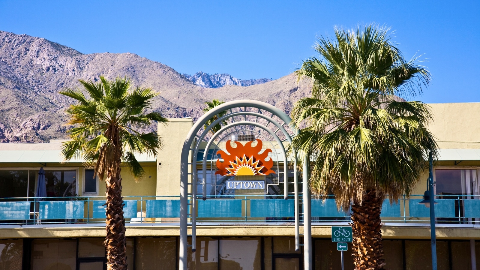 A photograph capturing the entrance to the uptown area of Palm Springs, California, with the prominent backdrop of the Mount San Jacinto Mountains. The image showcases the street leading into the uptown district, with commercial establishments lining the road. In the background, the Mount San Jacinto Mountains rise, creating a scenic backdrop against the clear sky.