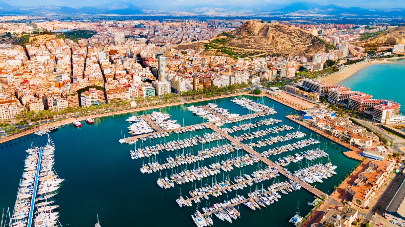 An aerial panoramic view of Alicante city port in the Valencia region, Spain. The image captures boats and yachts docked in the port, presenting a comprehensive overview of maritime activities. The cityscape surrounds the port area, featuring urban infrastructure against a clear backdrop.