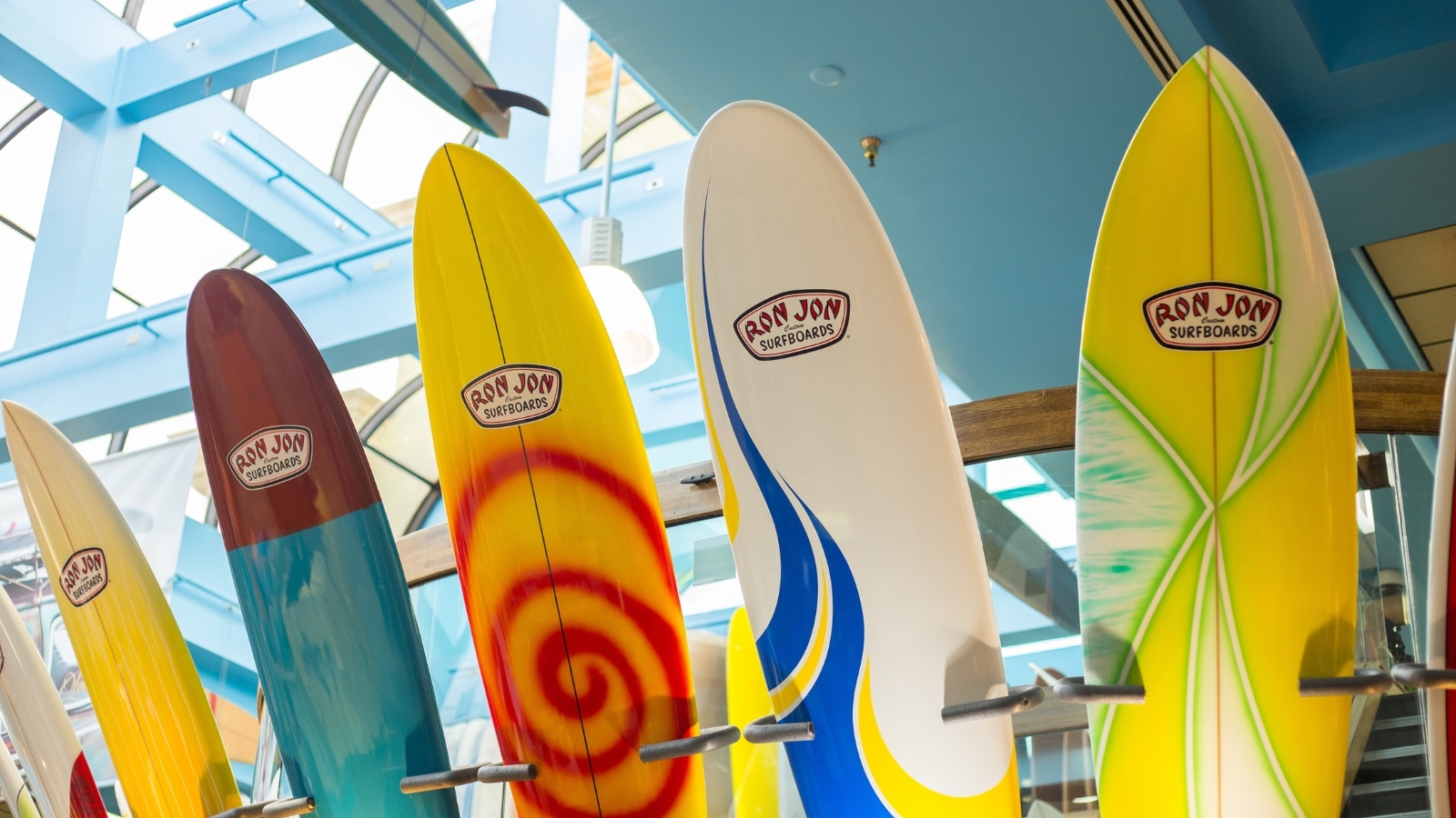 A close-up view of a row of colored surfboards arranged in a stand indoors. The image focuses on the surfboards' various hues and designs, presenting a visually distinct array.