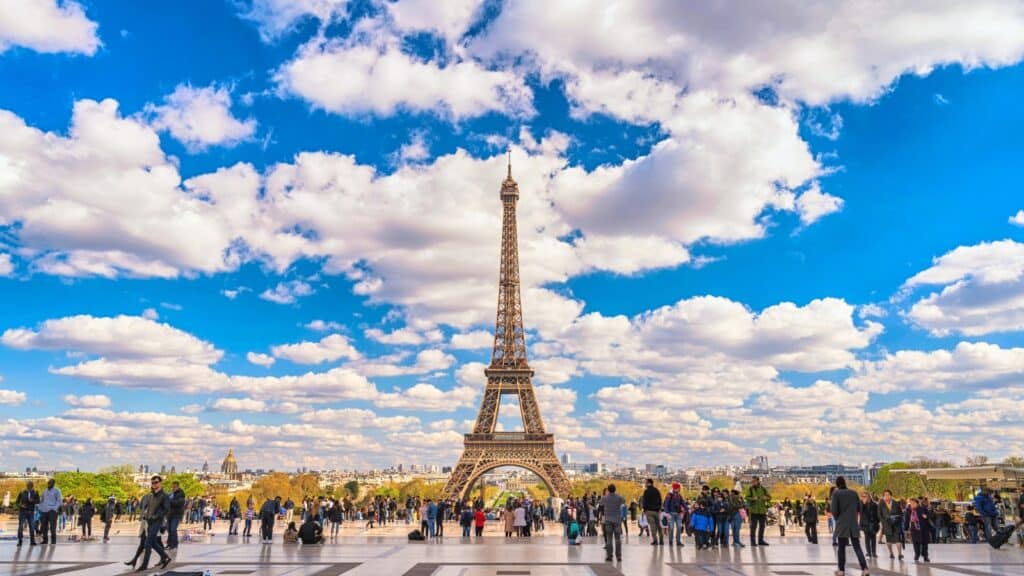 A photograph capturing the Eiffel Tower in the distance, with many people visible in the foreground. The image features a backdrop of beautiful clouds in the sky, adding to the scenic view.