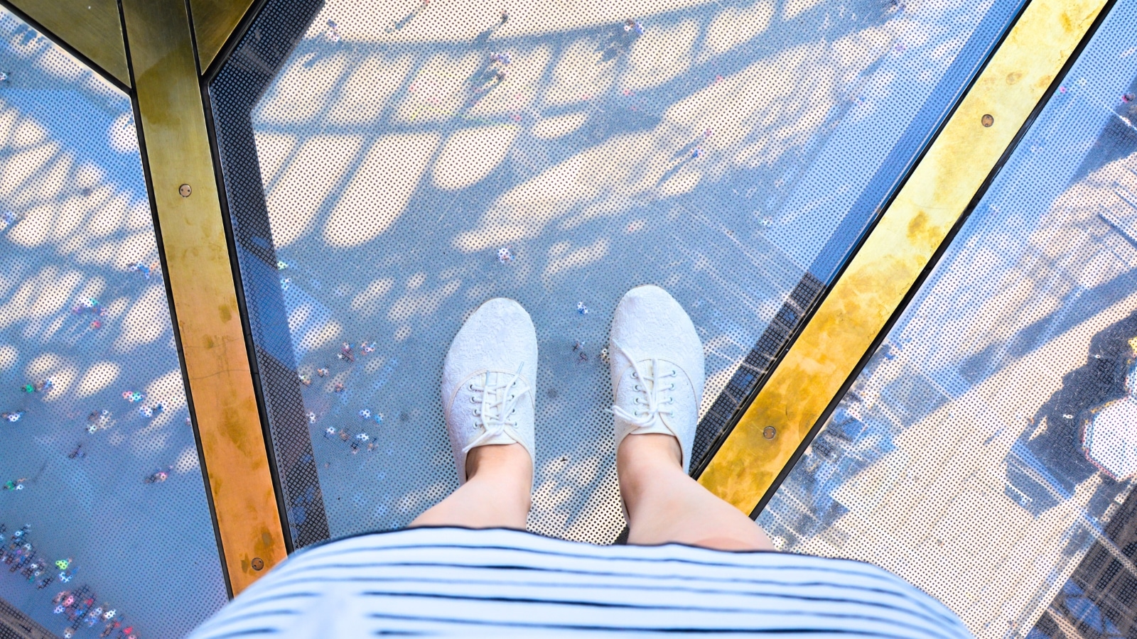 A photograph capturing the lower half of a person's body wearing white sneakers and a white skirt, taken from above on a glass floor at the Eiffel Tower.