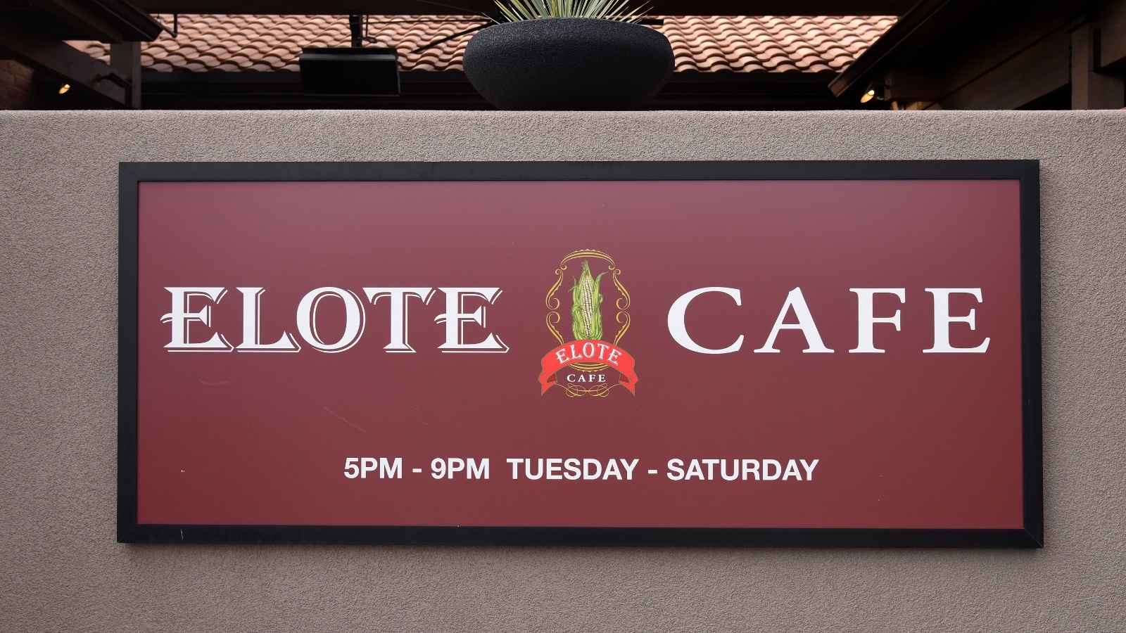 A close-up photograph of a red-burgundy sign displaying the inscription 'elote cafe' along with the establishment's schedule. The sign is prominently featured against a neutral background. The text is clearly visible and legible, providing information about the cafe's name and operating hours.