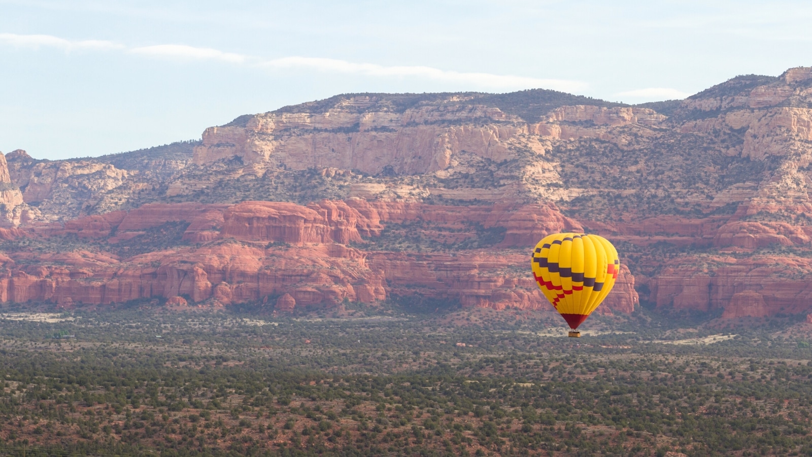 A photograph depicting a yellow hot air balloon floating above an arid area with rocky terrain in the background. The image captures the balloon's vibrant color against the neutral tones of the desert landscape.