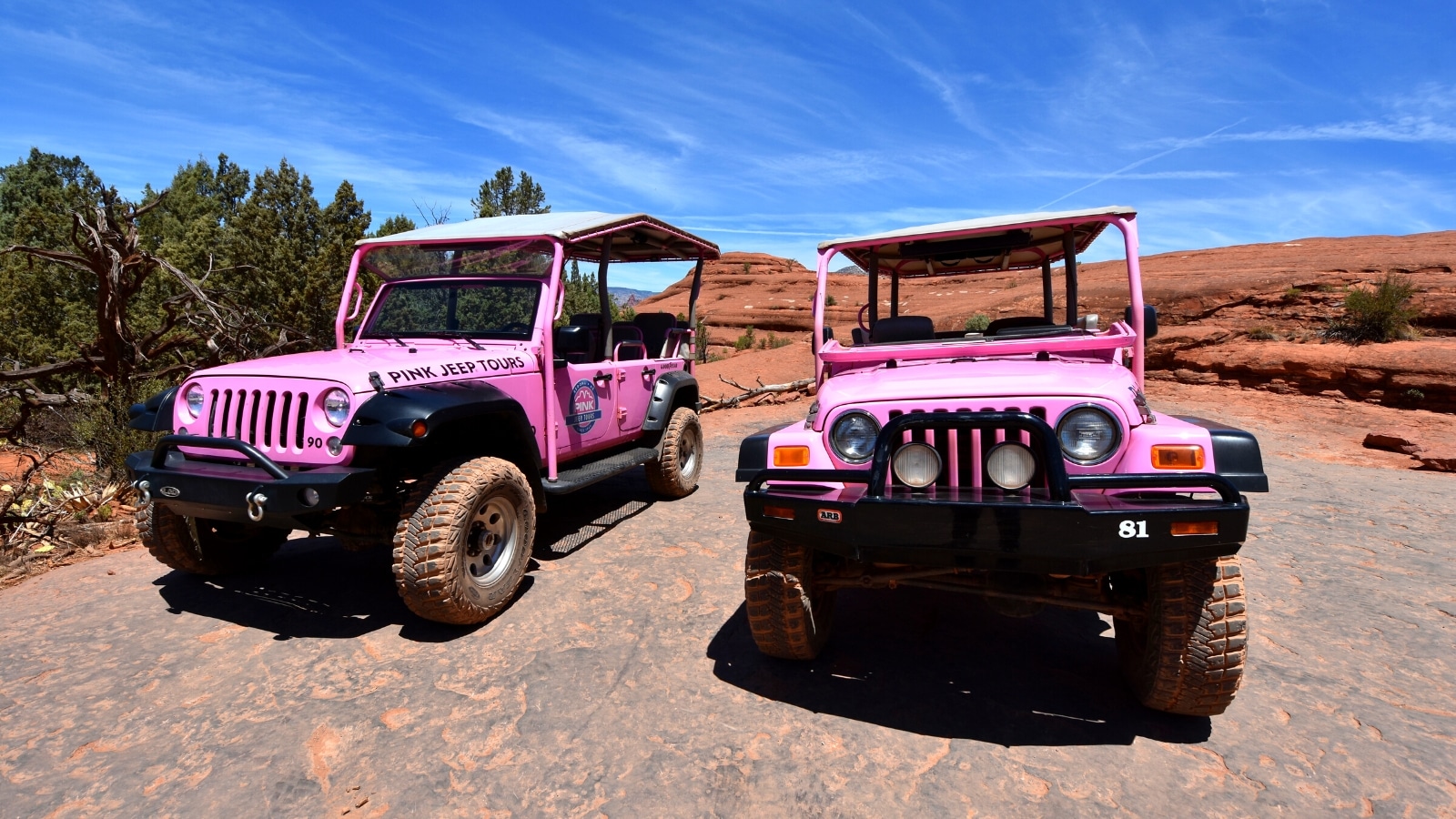 A photograph depicting two pink Jeeps traversing arid terrain in Sedona, Arizona. The image showcases the rugged desert landscape with minimal vegetation and rocky formations.