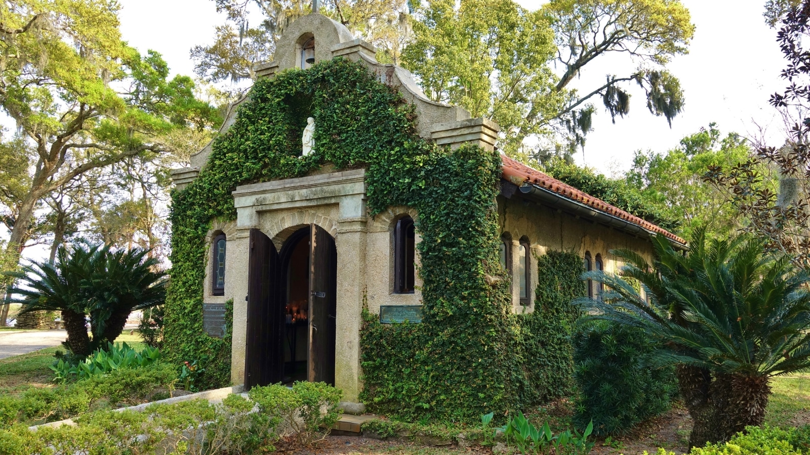 The image features a view of La Leche at Mission Nombre de Dios in St. Augustine, USA. The scene shows the historical architecture of the mission building, with its distinctive Spanish colonial style. Surrounding the mission are well-maintained gardens and pathways.