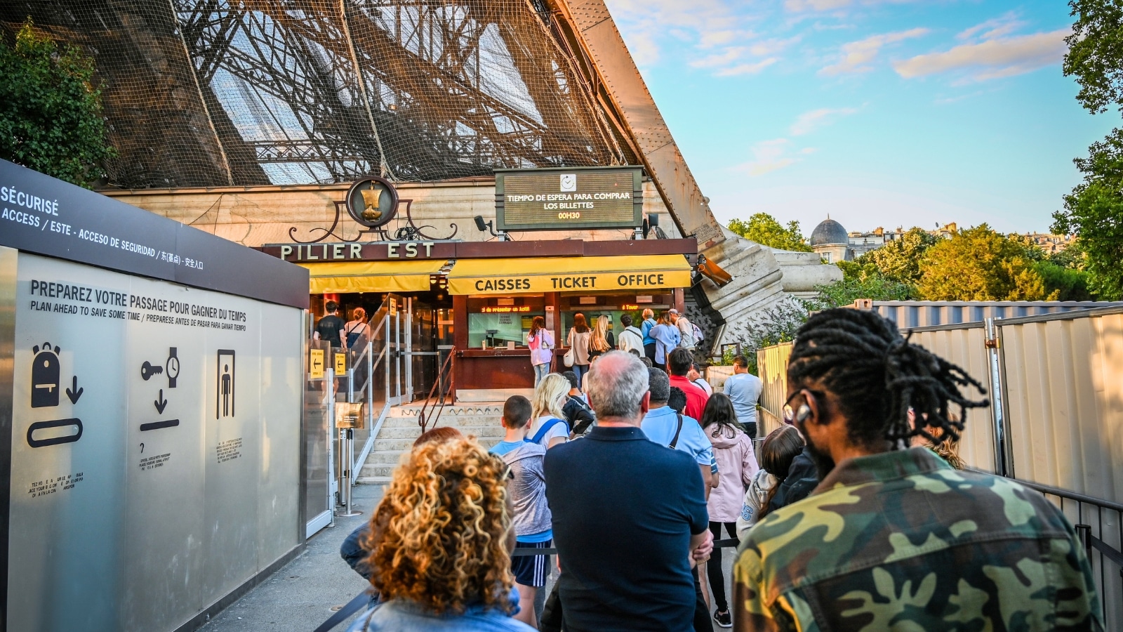 A photograph depicting people queuing to purchase tickets and access the Eiffel Tower in Paris. The scene portrays a typical day at the Eiffel Tower, with individuals of diverse backgrounds waiting for their turn to ascend the structure.