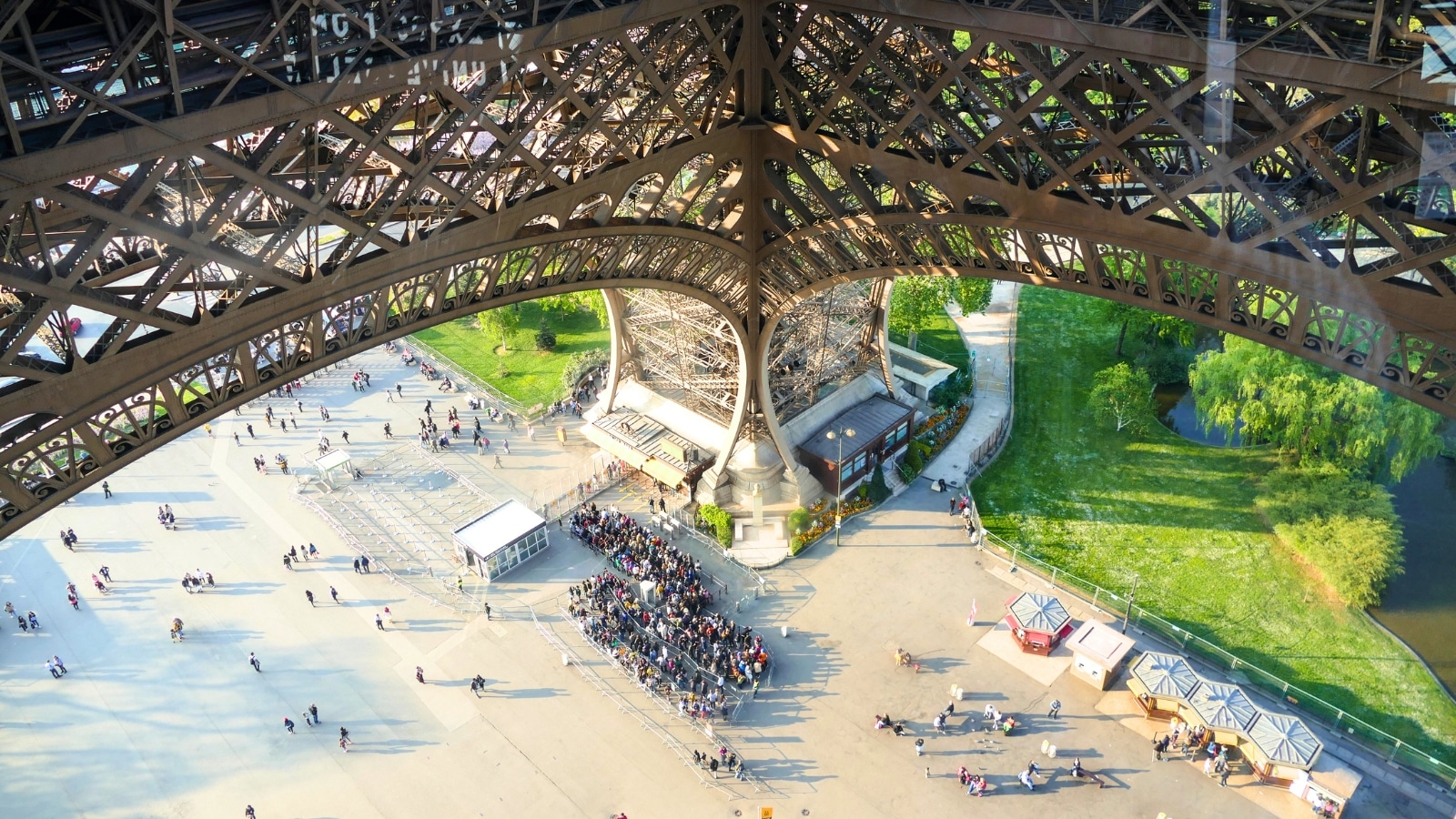 A top-to-bottom photograph capturing the entry line to the Eiffel Tower in Paris, France. The image shows a queue of people waiting to access the iconic landmark, with a pathway leading towards the base of the tower.