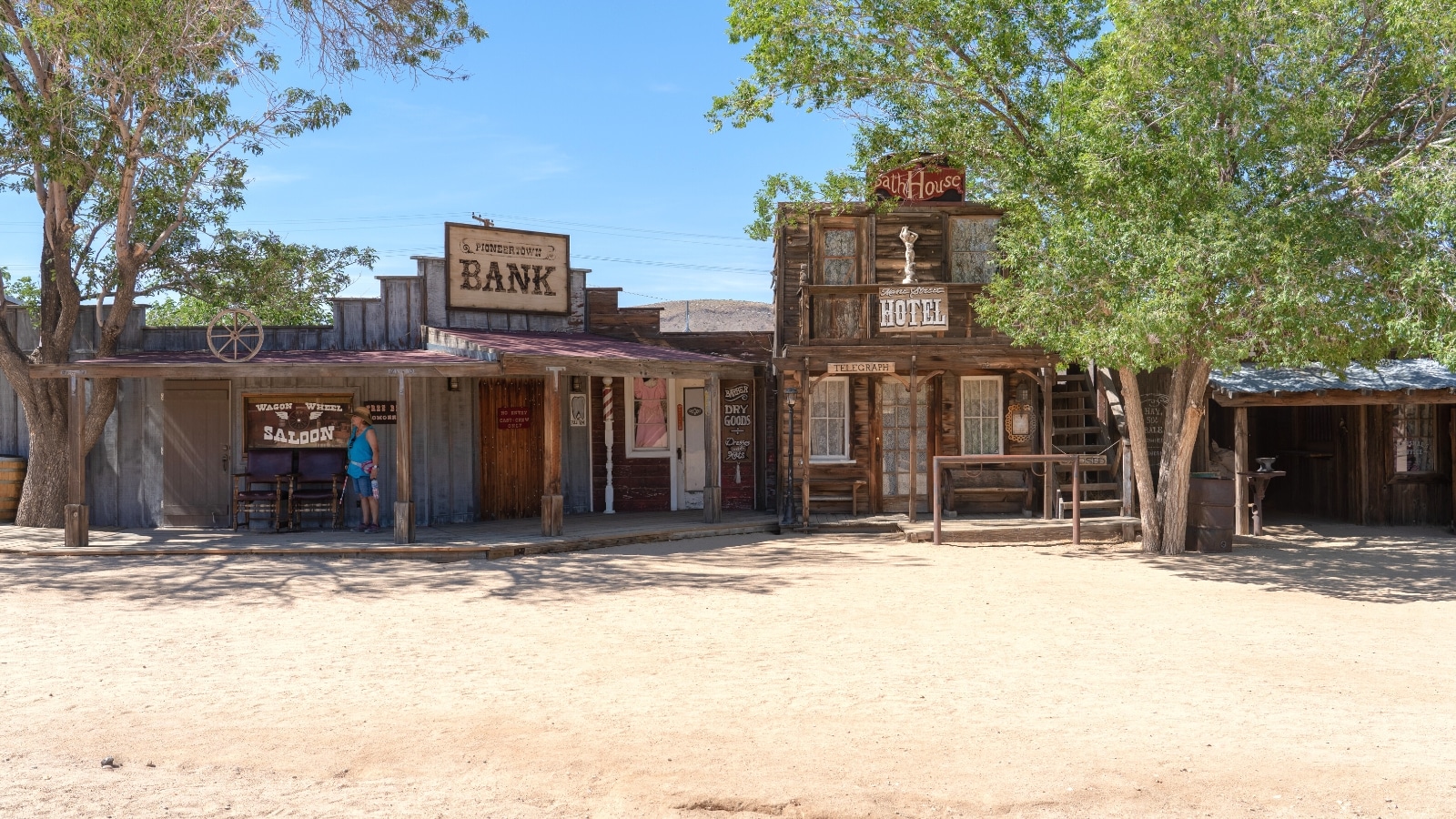 A photograph capturing the exterior of the Bank and Bath house in Pioneertown, California, as viewed from Mane Street. The image features the architectural elements of the structures, displaying a modest representation of the historical design prevalent in the region. The subdued colors and simple facades contribute to the overall aesthetic of the buildings.