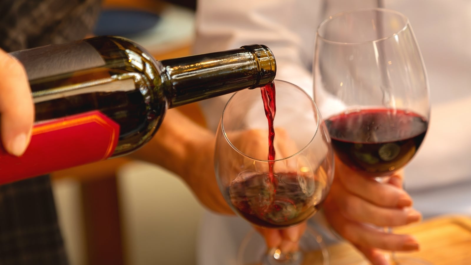 A photograph depicting the process of filling two glasses with red wine from a bottle. The image captures the fluid motion of the wine being poured into the glasses, showcasing the rich color and texture of the wine.