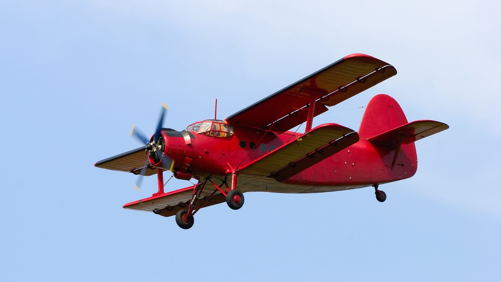 A photograph capturing a red airplane with double wings and a front-mounted propeller against a clear blue sky. The image provides a straightforward depiction of the aircraft's design, featuring distinctive characteristics such as the red color, double wings, and visible propeller.