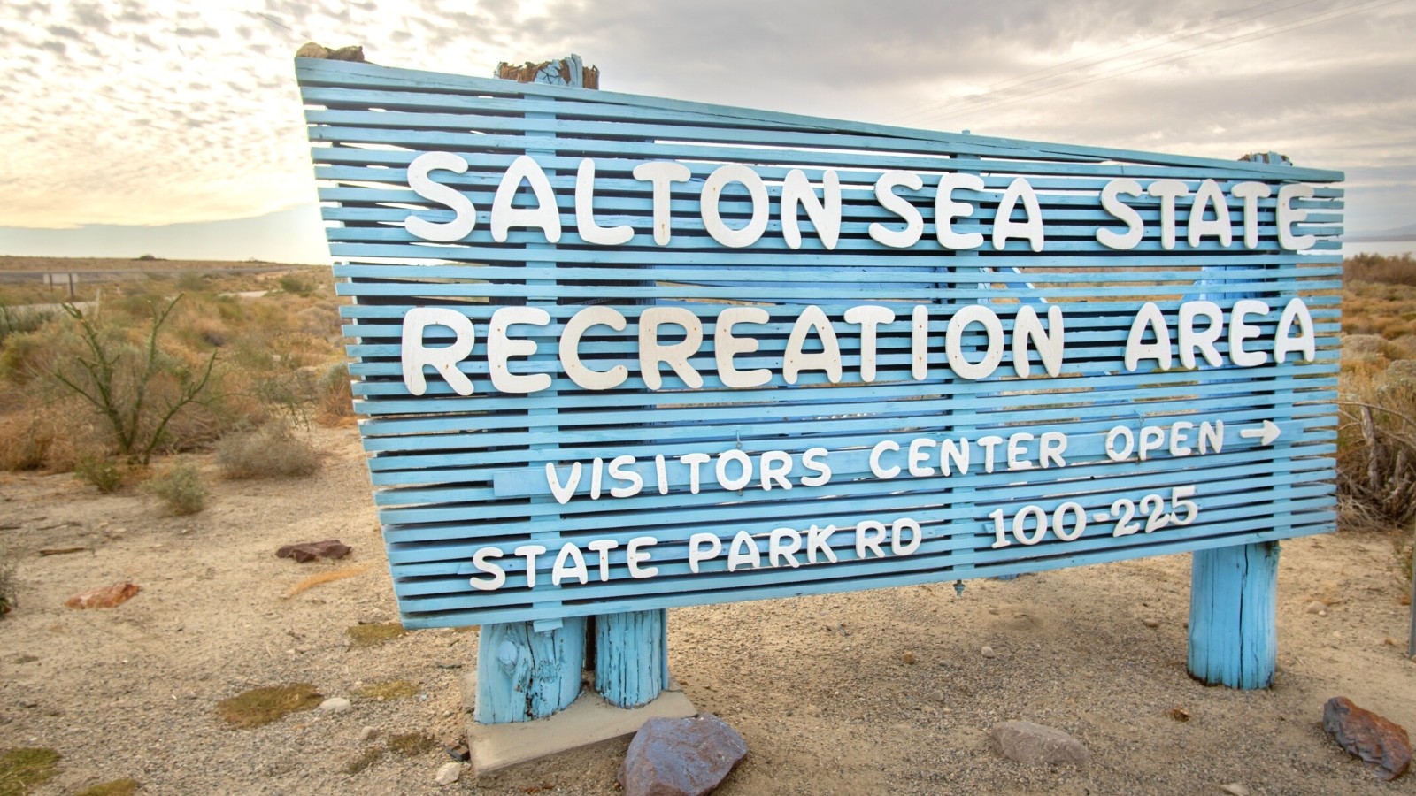 A photograph capturing the entrance sign to Salton Sea State Recreation Area. The sign features a sky-blue background with white text indicating the name of the recreational area. The surrounding landscape consists of a desert area against a cloudy sky.