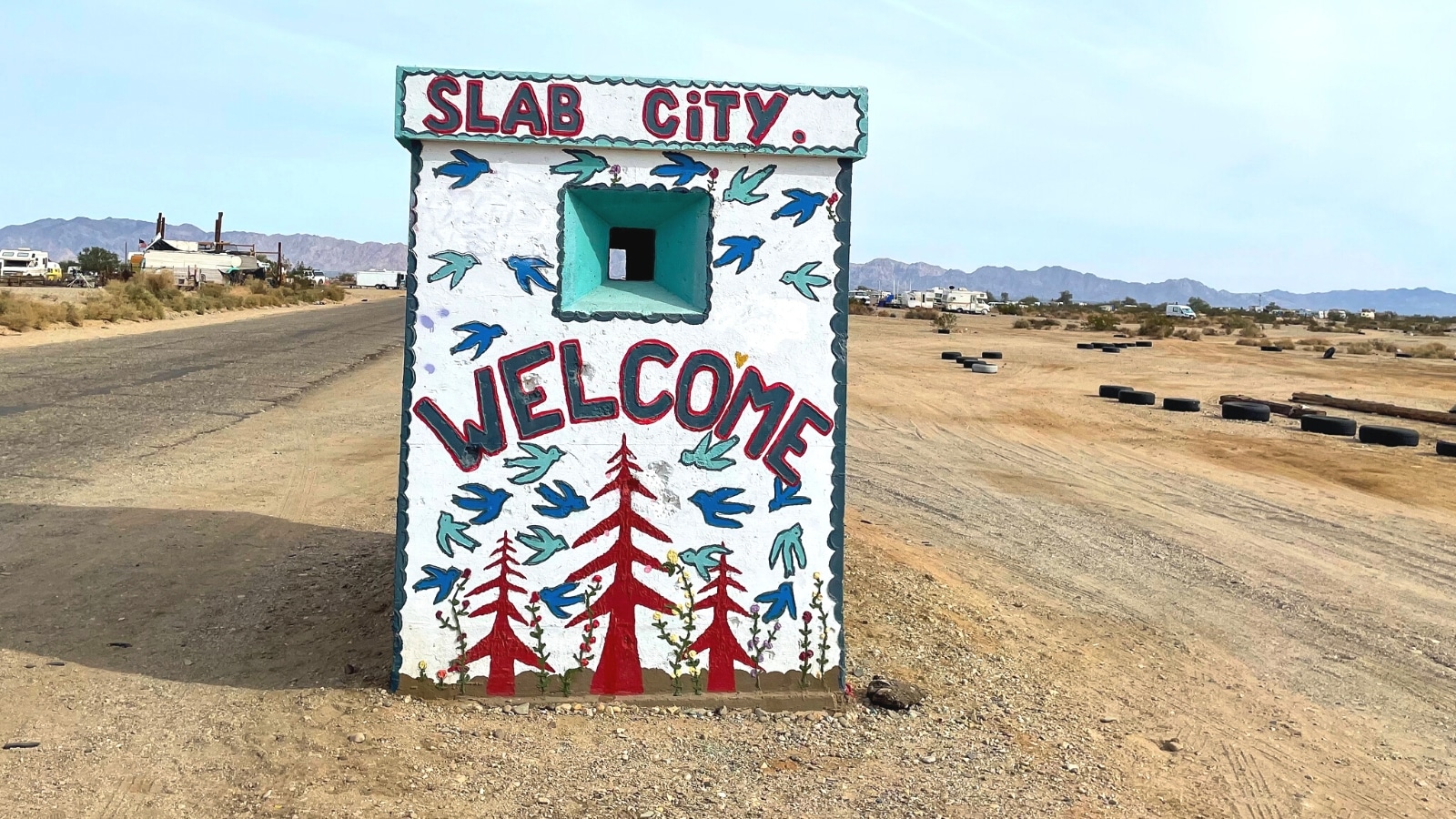 A photograph depicting a small square building in Slab City, California, standing in an arid area. The building features the words 'Slab City Welcome' prominently displayed.