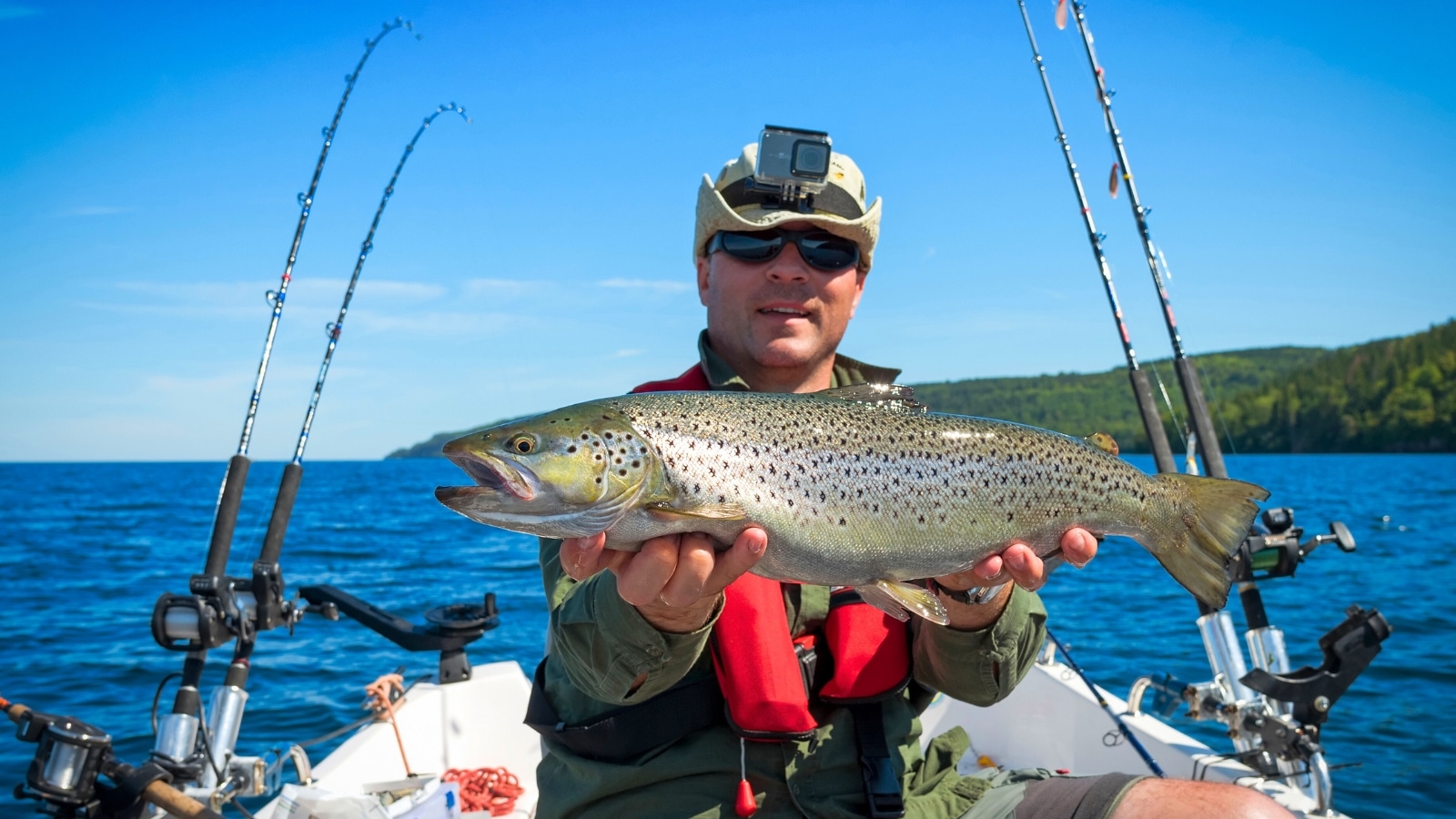 The image shows a St. Augustine fisherman holding a trout up close while on a boat in the water. Surrounding the fisherman are numerous fishing rods placed on both sides of the boat.