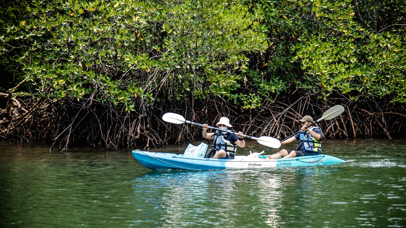 The image features two men sailing in a kayak, each holding oars in their hands. The background is filled with dense bushes, creating a natural and secluded setting.