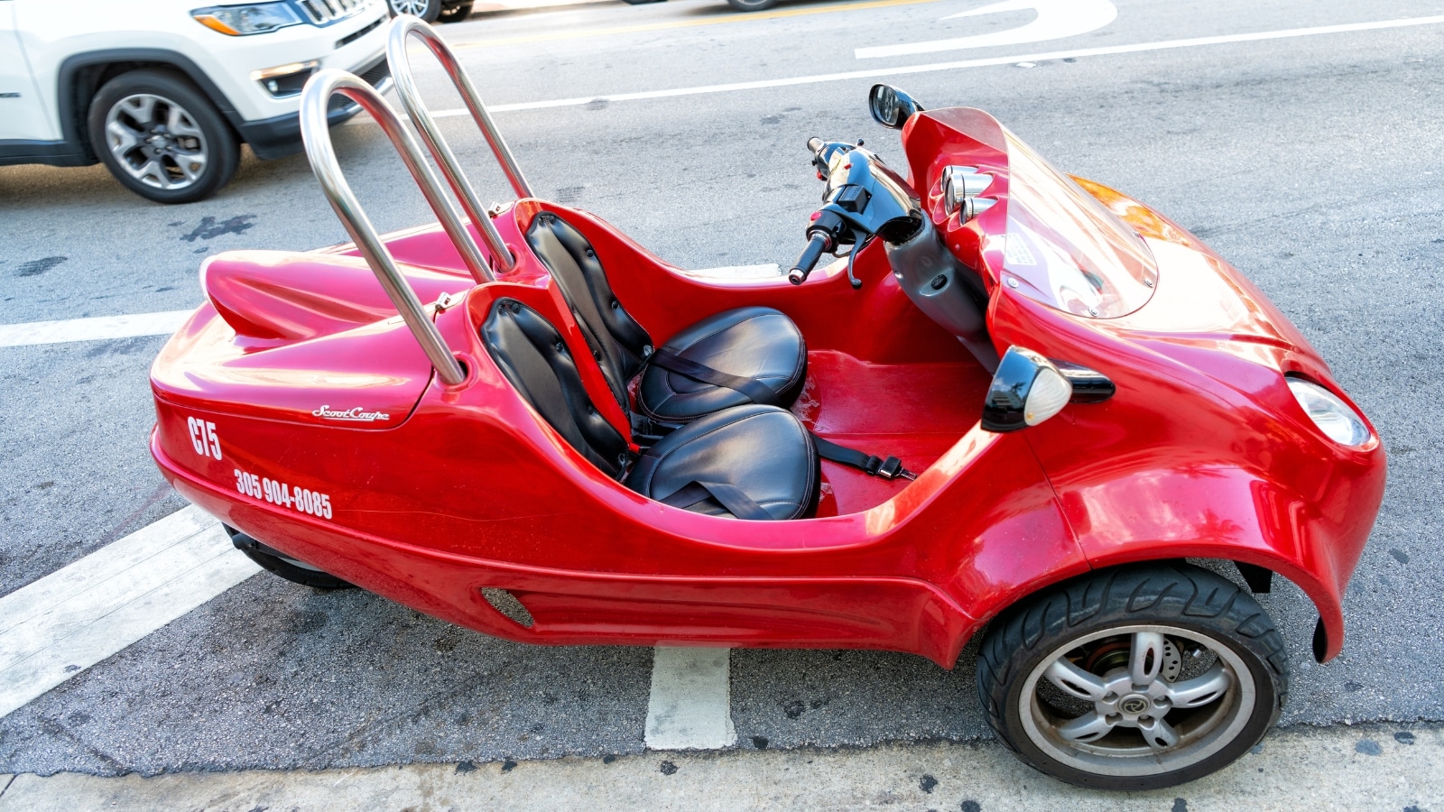 The image displays a St. Augustine ScootCoupe, a red three-wheeled car designed with two seats, positioned on a standard road.