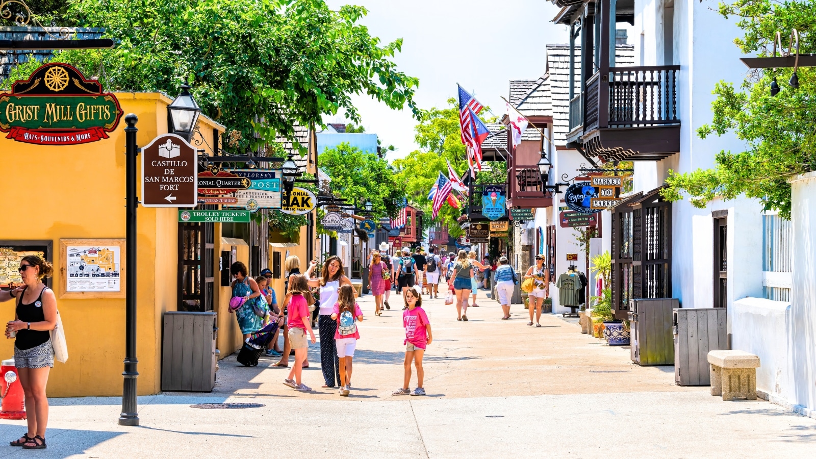 The image captures a scene in St. Augustine, USA, where people are walking and shopping along St. George Street on a summer day. The street is lined with stores, shops, and restaurants typical of the old town Colonial quarter.