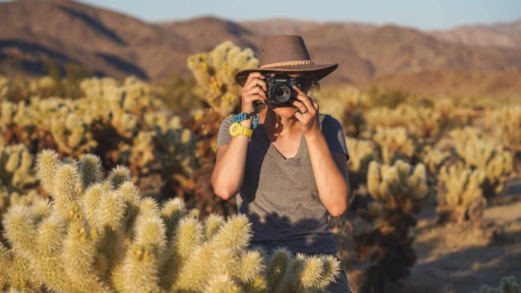 Photo of a girl taking a photo in Joshua tree national park. She is dressed in a gray T-shirt and a straw hat, holding a black camera in her hands and covering her face with it. There is a yellow watch on her wrist. She stands surrounded by Teddy-bear cholla plants.