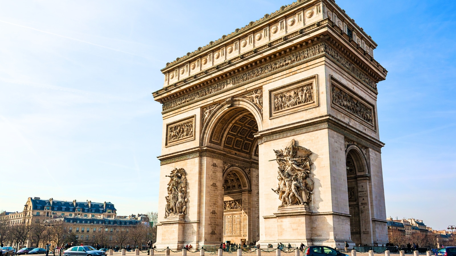 The image captures a sunny view of the Arc de Triomphe in the Étoile Square in Paris, France. The iconic monument stands prominently in the center of the square. Surrounding the Arc de Triomphe are wide avenues and bustling city streets.