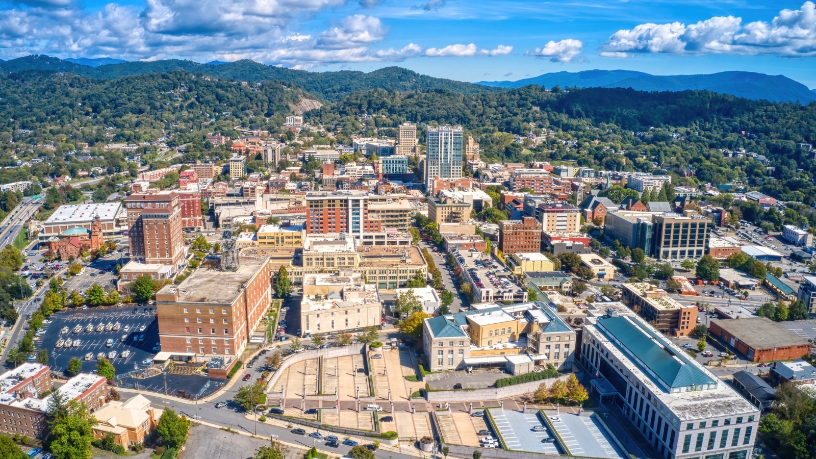 The image provides an aerial view of Asheville, North Carolina, with mountains in the background and the cityscape in the foreground.