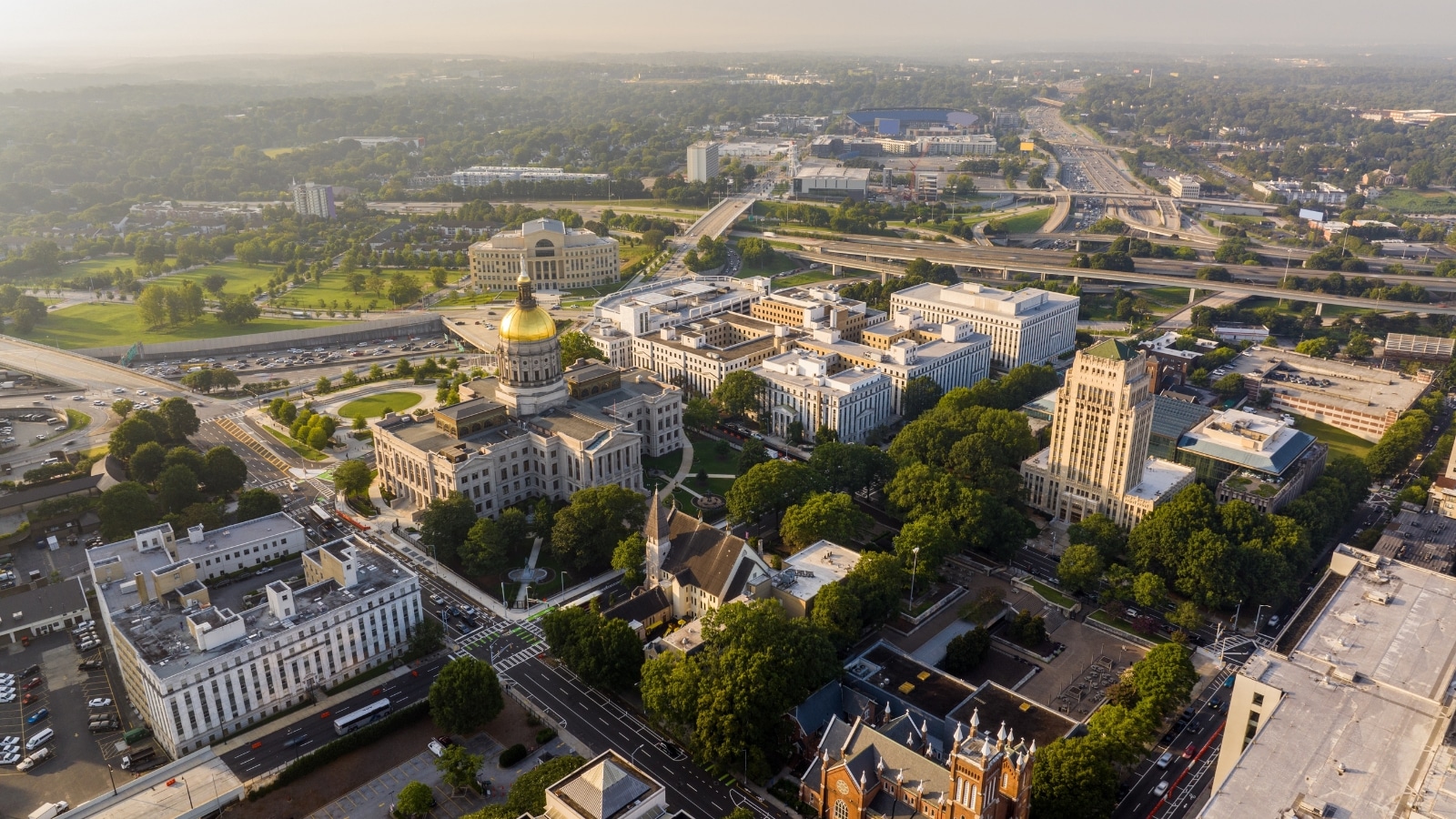 The image provides an aerial perspective of Georgia Capitol Hill and surrounding government buildings under an overcast sky. Along the streets, there is an abundance of greenery, contributing to the city's landscaping. The architecture of Capitol Hill and the government buildings is prominently displayed, showcasing the official and administrative hub of Georgia.