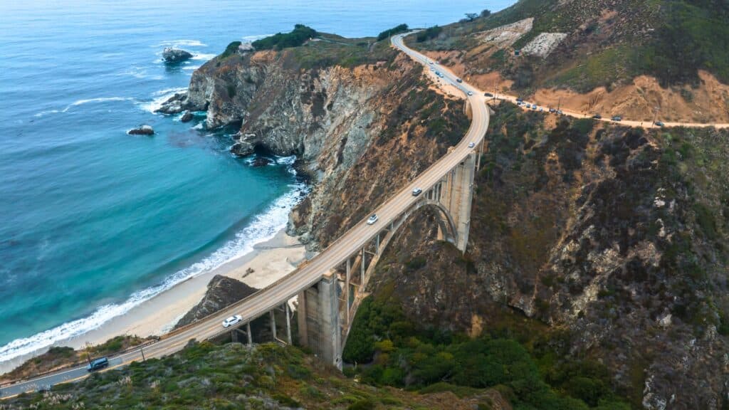 The image displays an aerial view of the Bixby Bridge in California, USA, showcasing its architectural features and the surrounding landscape. The bridge is positioned near the Pacific Ocean, adding to its scenic appeal.