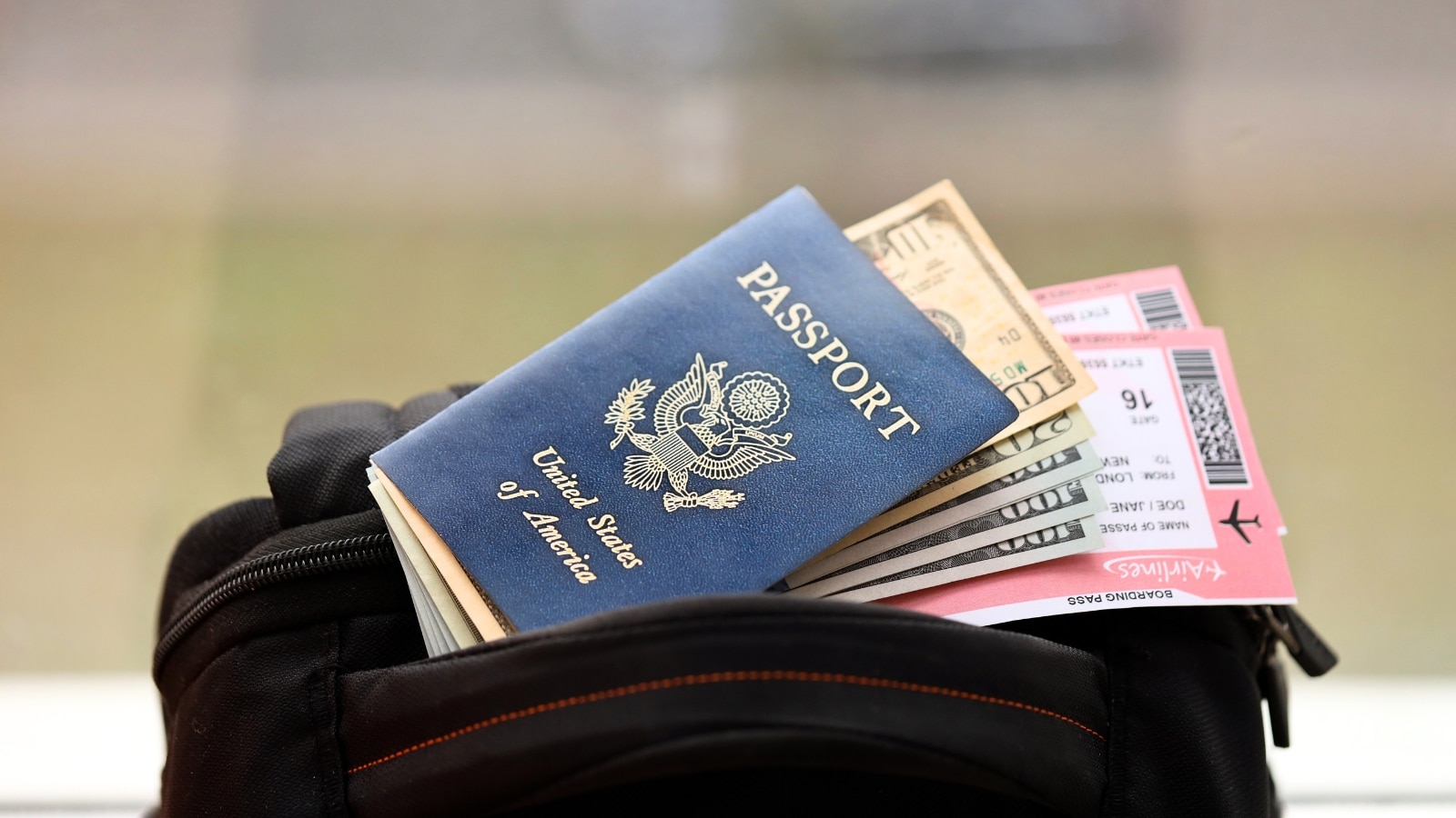 The image depicts a blue United States of America passport placed alongside money and airline tickets on top of a tourist backpack.