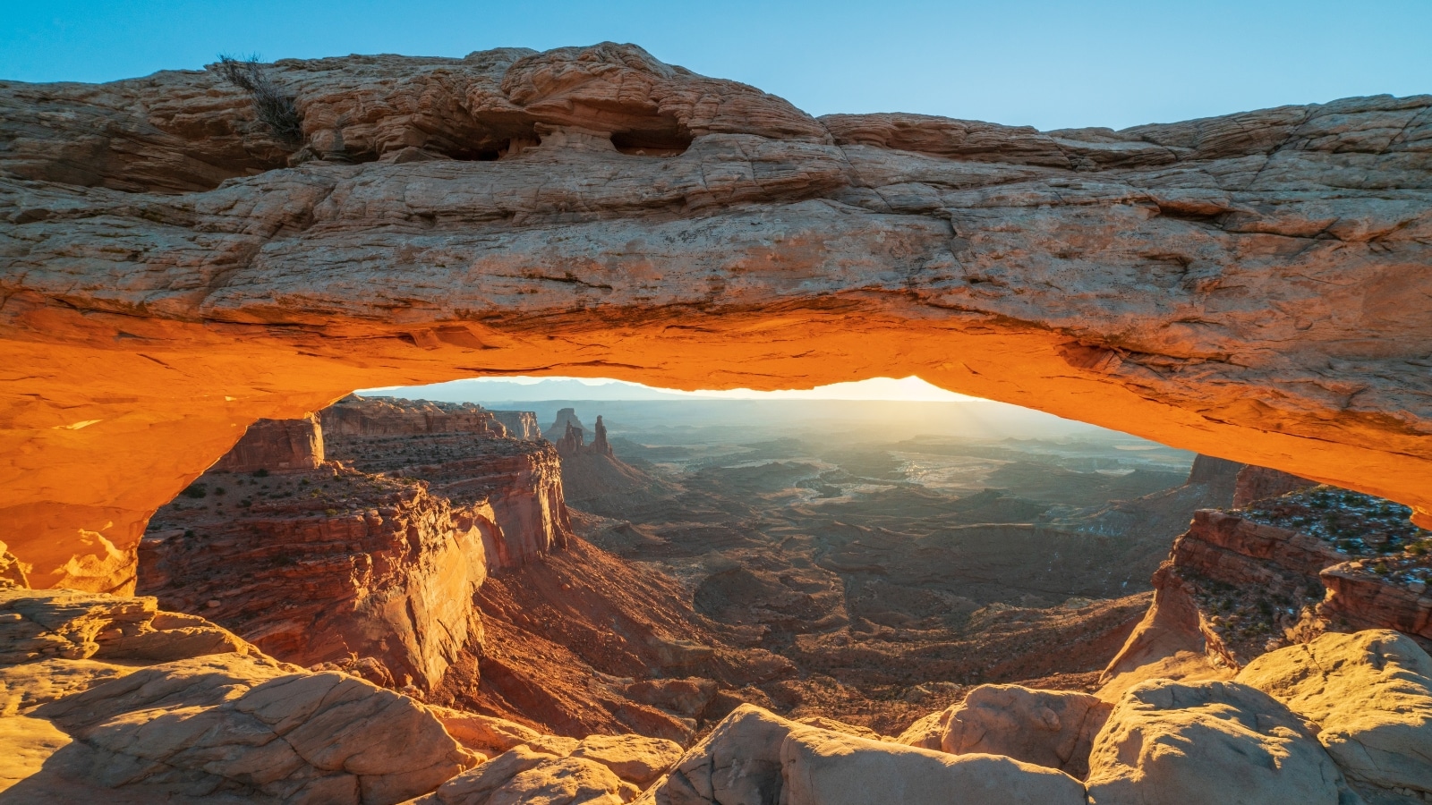 The image captures the iconic Mesa Arch at sunrise, situated in Canyonlands National Park in southeastern Utah, USA. The natural arch frames a breathtaking vista of rugged terrain and vast canyons, typical of the region's geological features.