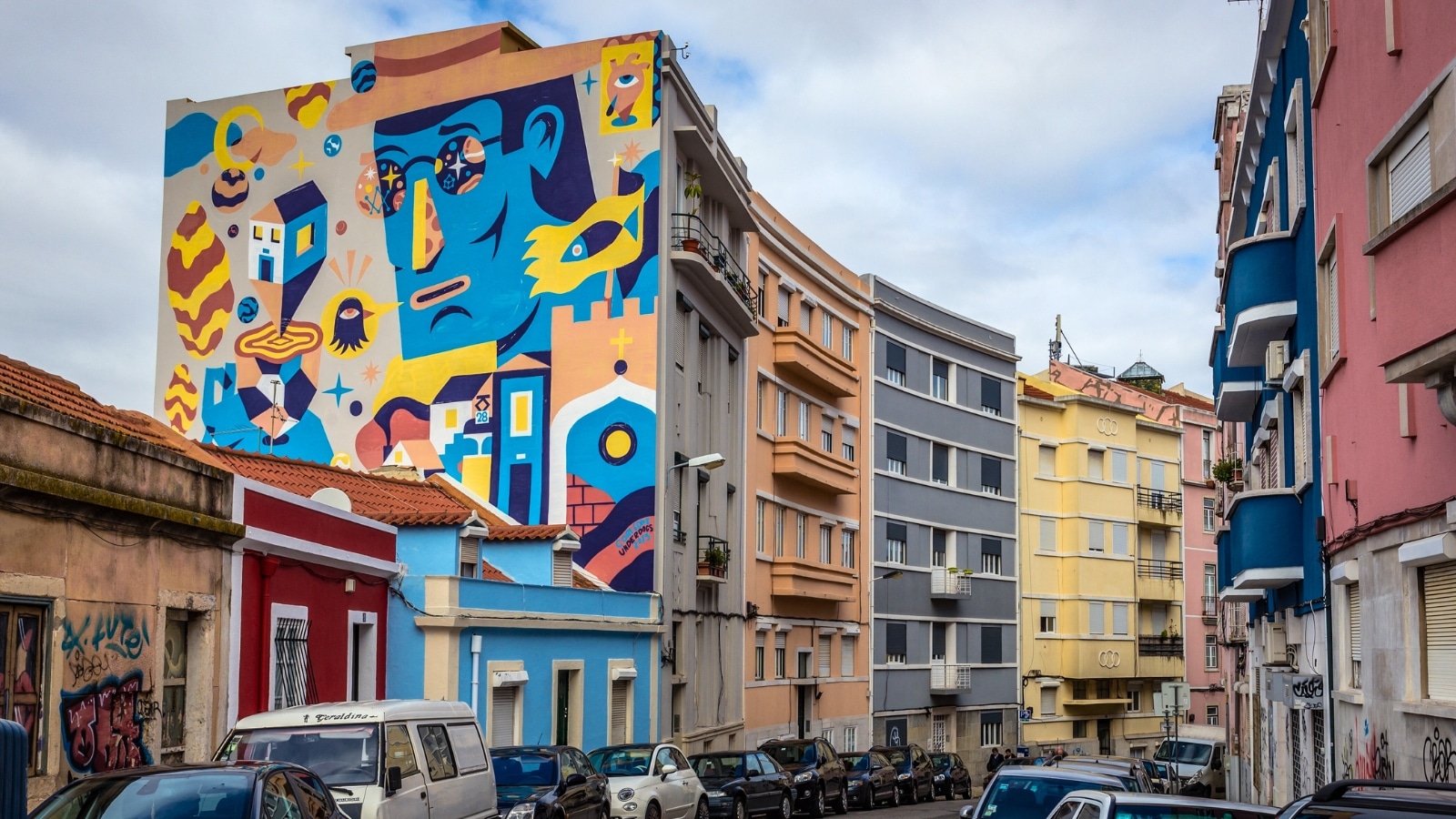 The image features Damasceno Monteiro street in the Graca area of Lisbon, Portugal. A prominent feature is a large colored graffiti covering the entire facade of a building along the street. Numerous cars are parked below, indicating urban congestion typical of city areas.