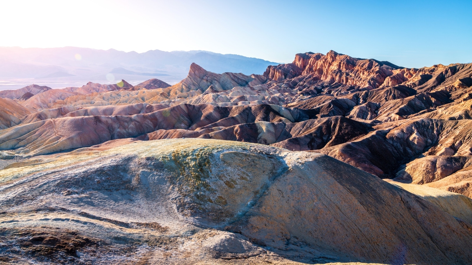 The image captures a scenic view of the badlands from Zabriskie Point in Death Valley National Park, California. The landscape features rugged terrain with distinctive geological formations, showcasing erosion patterns and unique color variations typical of badlands environments.