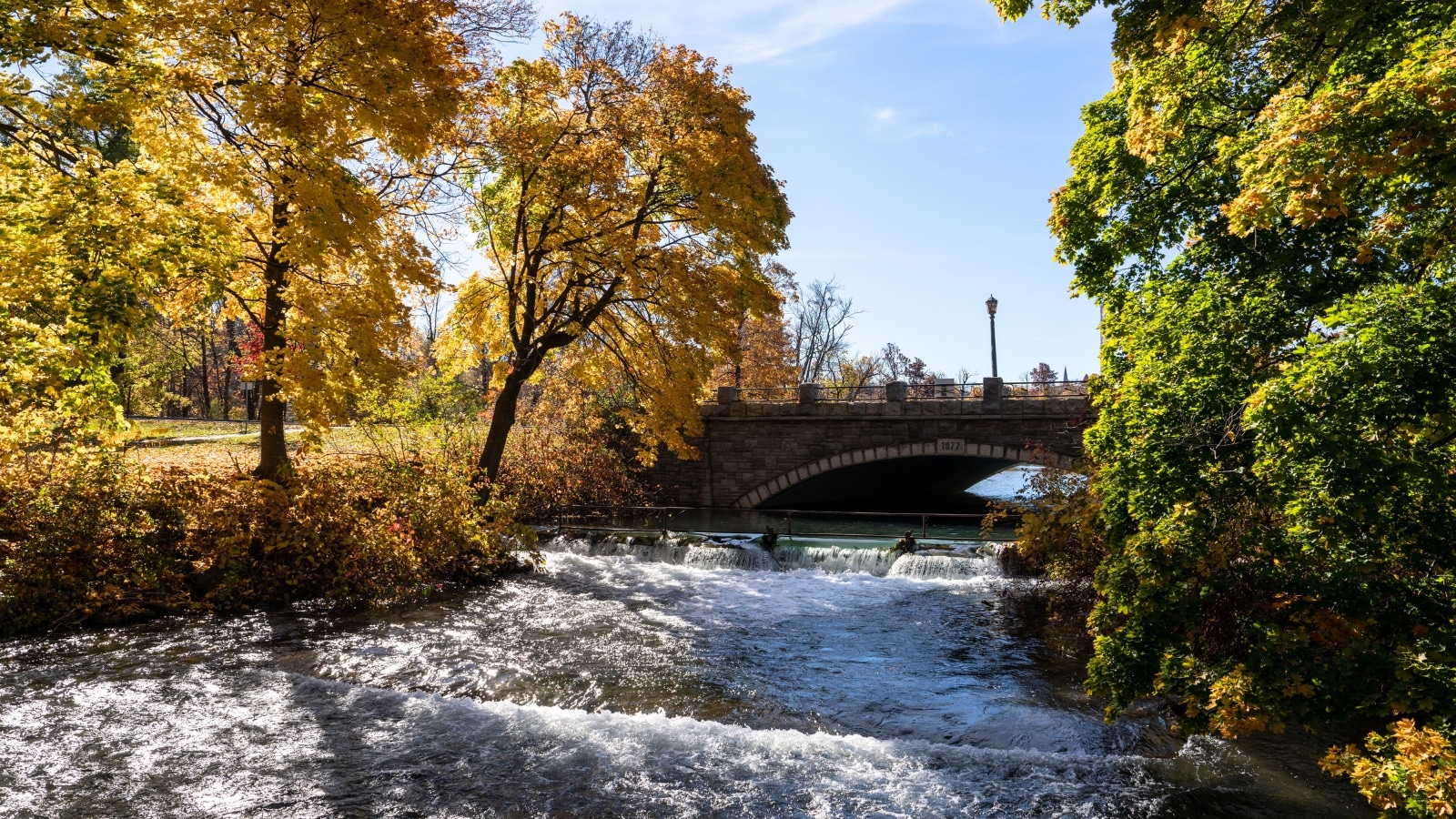 The image depicts a scenic view of the Dufferin Islands, showcasing a river, a bridge, and abundant greenery adorned with green and orange leaves. The bridge spans gracefully over the tranquil river, blending seamlessly with the natural landscape.