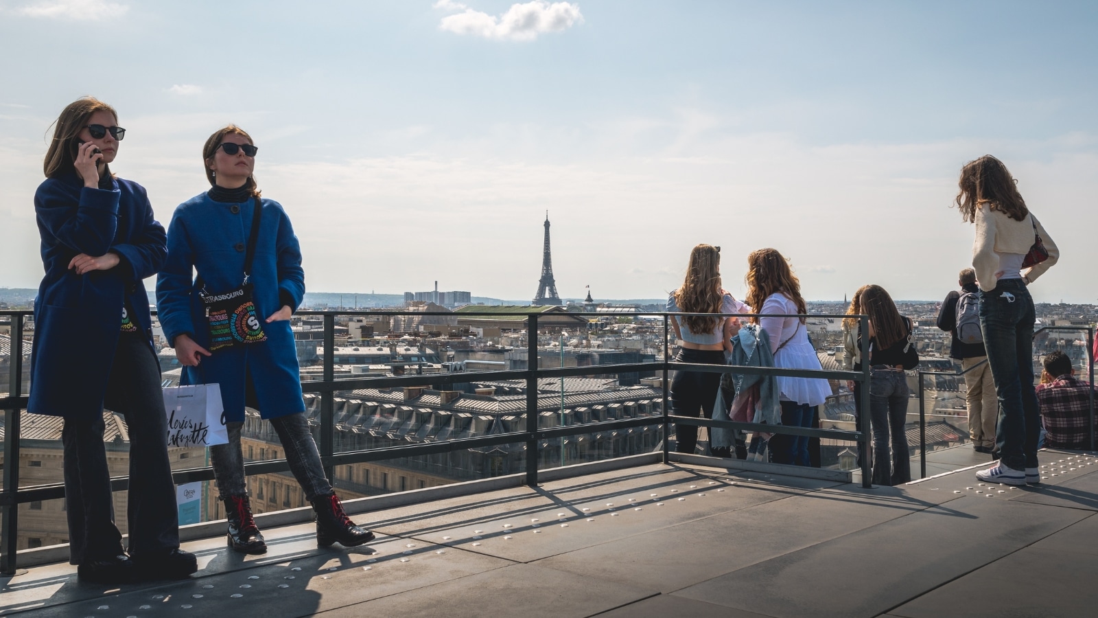 The image captures a panoramic view above the rooftops of Paris, showcasing the terrace from Galeries Lafayette with the iconic Eiffel Tower in the background. Approximately ten people can be seen standing on the observation deck, enjoying the scenic vista of the city. The observation deck offers a clear view of Paris's urban landscape.