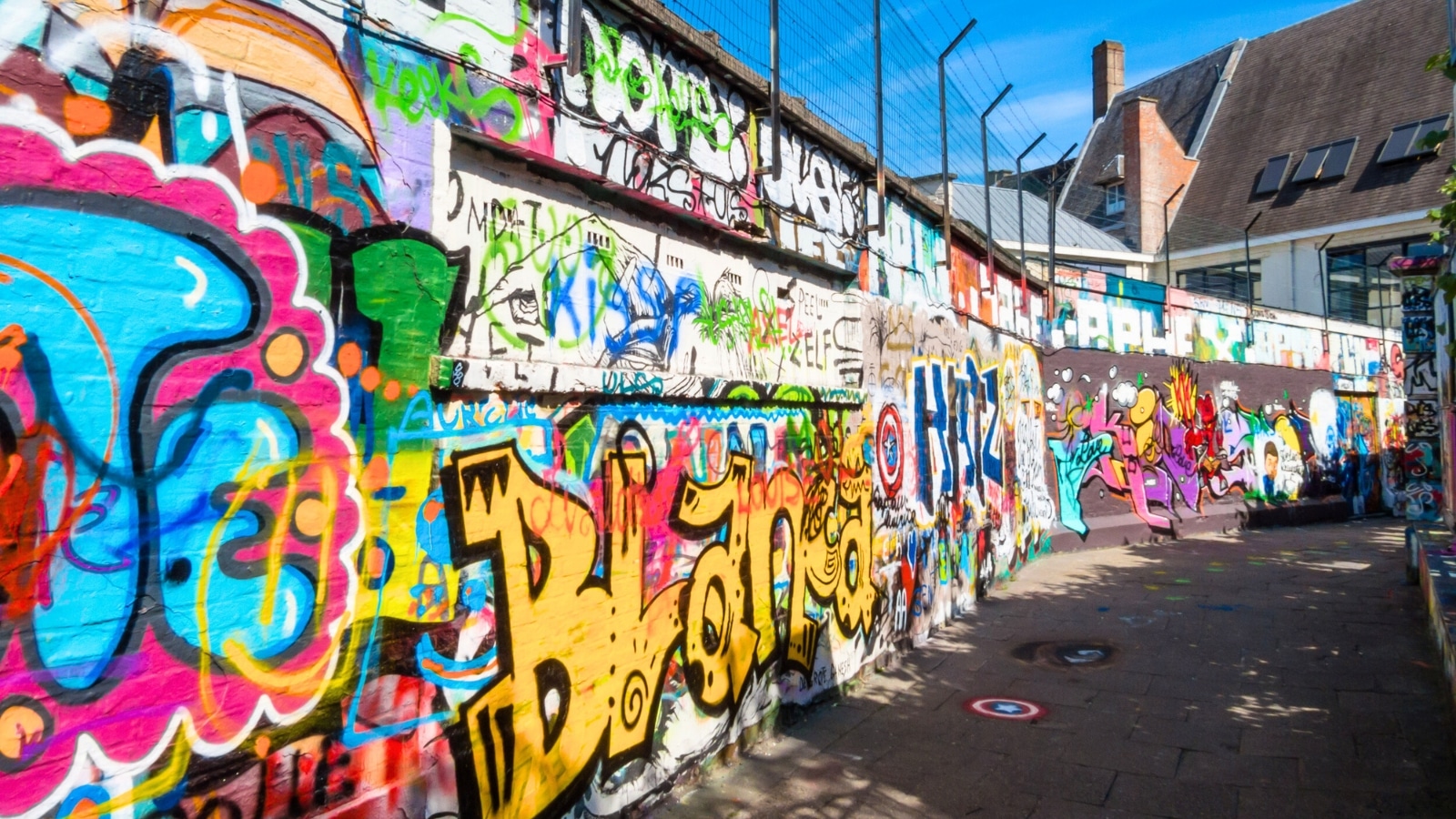 The image captures a scene from Graffiti Street (Werregarenstraat) in Ghent, Belgium. The street is known for its vibrant graffiti art, which covers the walls and surfaces of buildings along the thoroughfare. The graffiti displays a variety of styles, colors, and themes, showcasing the artistic diversity found in this area.