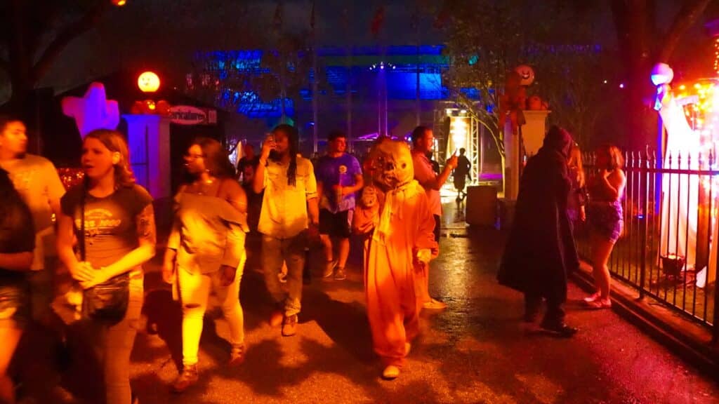Crowds of thrill-seekers traverse the park, immersed in the eerie ambiance. Masked figures adorned in terrifying costumes lurk in every corner, adding to the spine-tingling atmosphere. Neon signs flicker ominously, casting eerie shadows across the pathways.