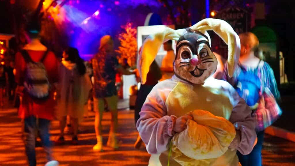 In the image depicting Halloween Horror Nights at Universal Studios Orlando, Florida, a gloomy atmosphere pervades the scene. The foreground showcases a close-up of a man dressed as a somber hare, adding an eerie touch to the setting.