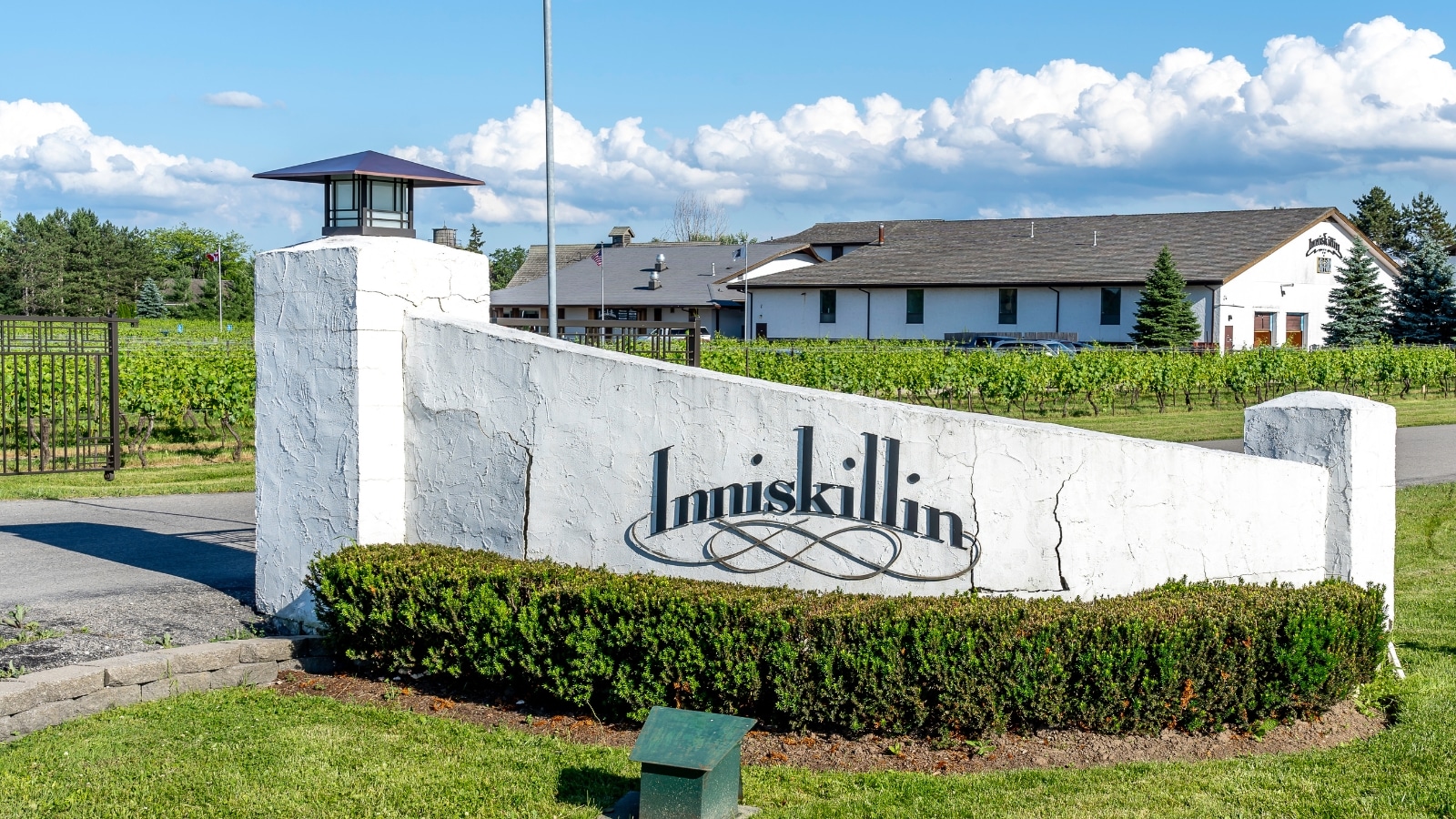 The image captures the entrance to Inniskillin Wines Winery in Niagara-on-the-Lake. The entrance features a sign with the inscription "Inniskillin," marking the beginning of the winery's premises. In the background, sprawling fields of vineyards are visible, typical of the winery's scenic location. A one-story house with white walls and a triangular roof complements the rural setting.