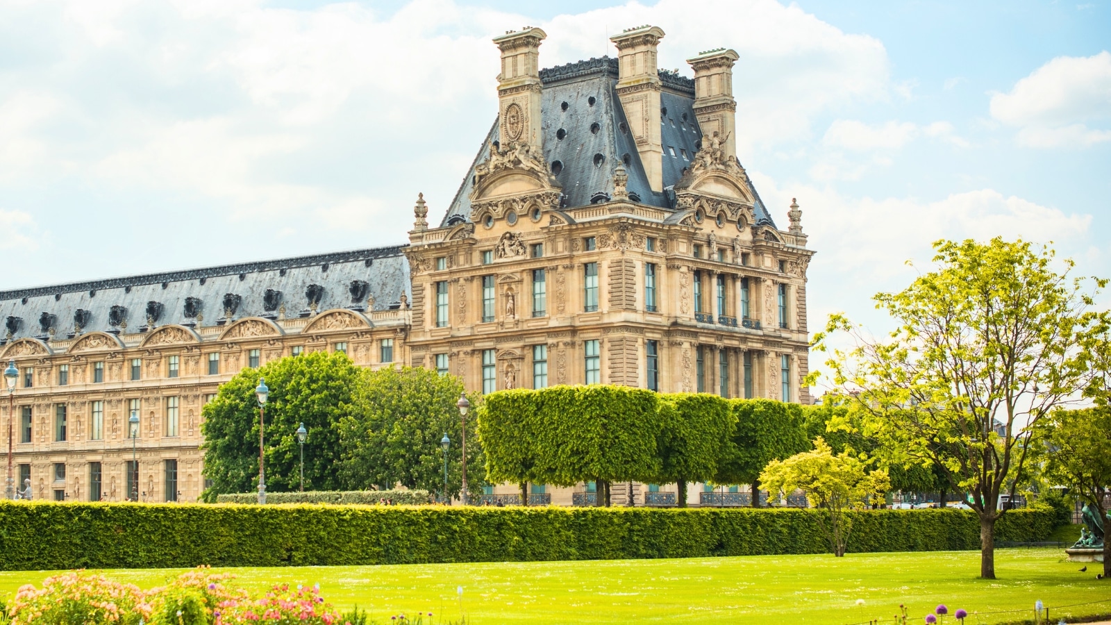 Jardin des Tuileries in Paris, France. The foreground features well-groomed lawns, meticulously trimmed bushes and trees, and squared-off shrubbery. In the background, there is a prominent large historical building, adding a sense of grandeur and historical significance to the scene.