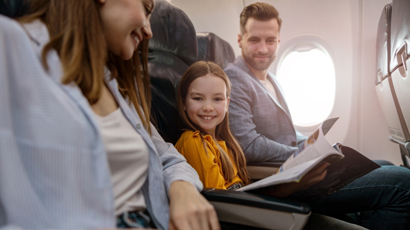 The image depicts a joyful little girl traveling with her parents on an airplane. The composition shows the mother seated on the left side, the daughter in the center, and the father on the right.