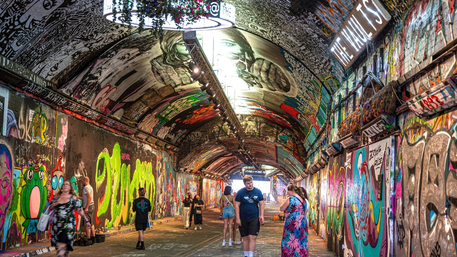 The image features Leake Street Tunnel in London, known for its extensive graffiti art. The tunnel is adorned with a plethora of colorful graffiti artworks, showcasing a vibrant and artistic atmosphere. In the scene, people can be seen walking through the tunnel, observing and appreciating the graffiti on display.