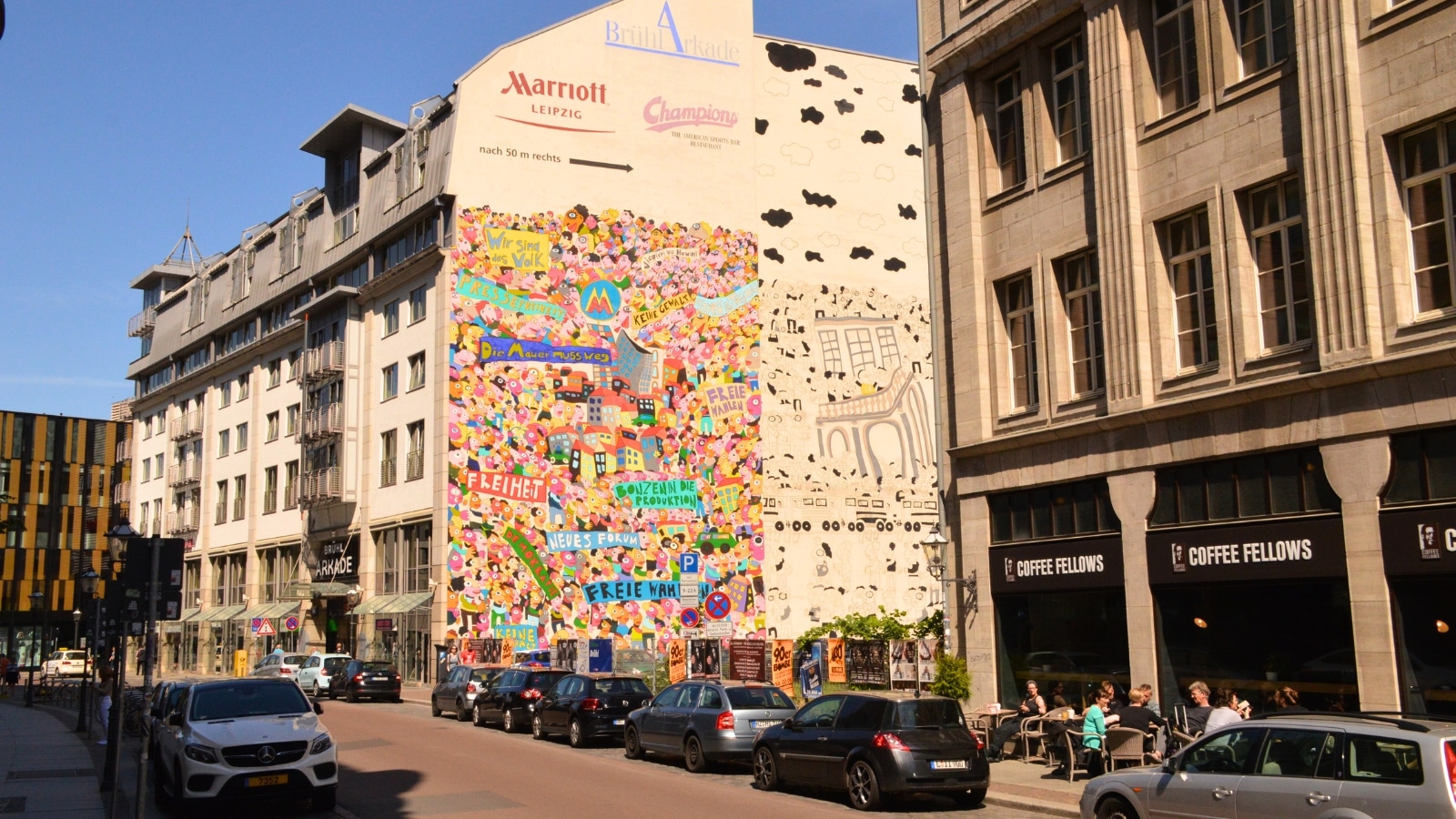 The image shows a piece of large colored graffiti on the wall of a house in Leipzig, Germany. Parked cars are visible below the graffiti, adding to the urban setting of the scene.