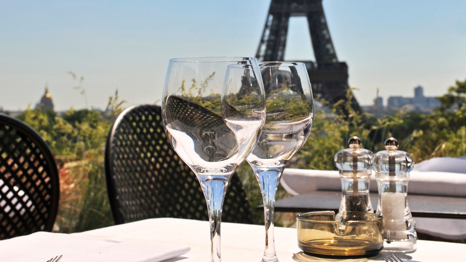 The image depicts a table in a restaurant setting with two glasses of white wine, positioned to overlook the Eiffel Tower.