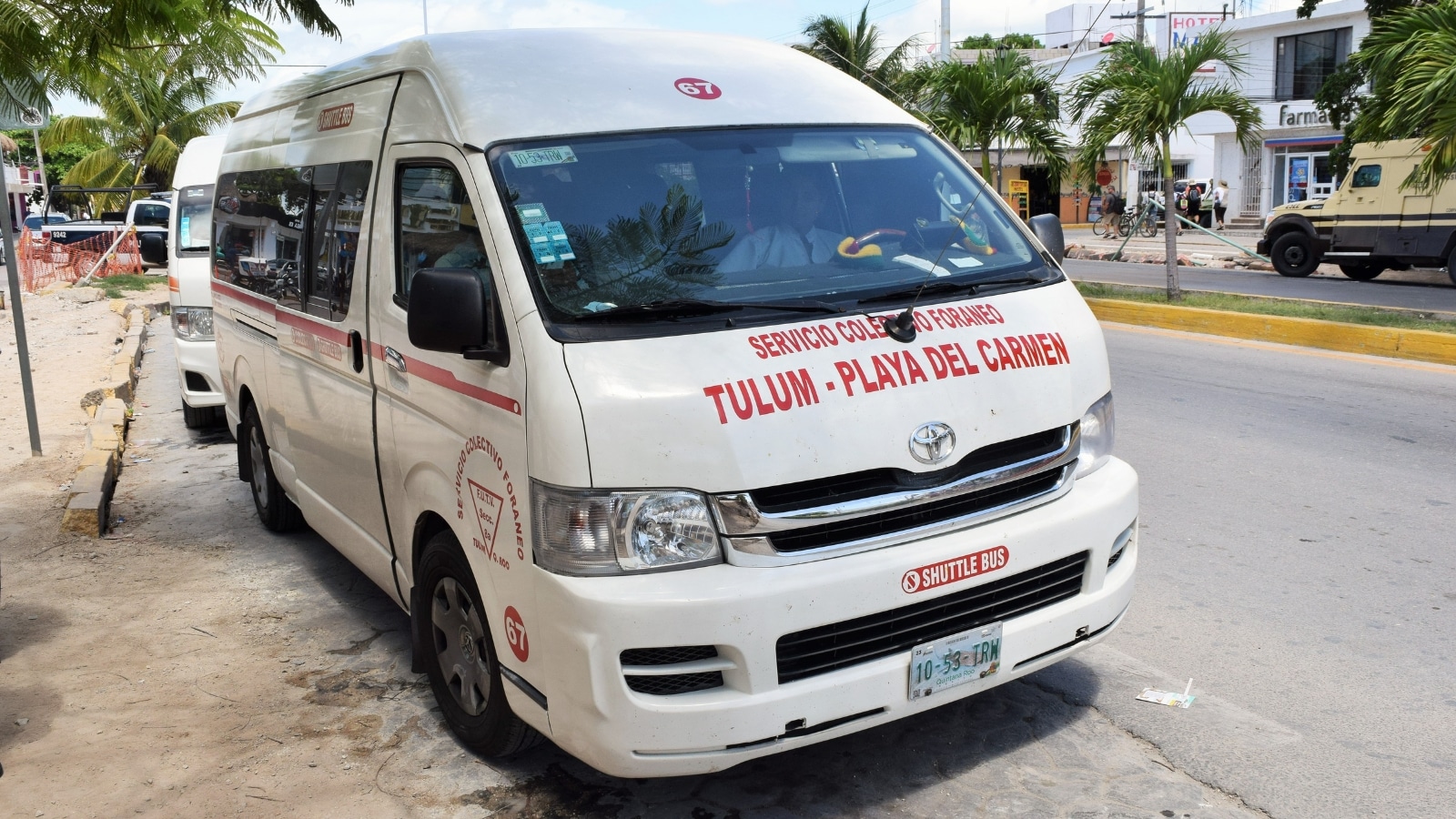 The image shows a close-up view of a white mini bus traveling from Tulum to Playa del Carmen. The bus is adorned with red inscriptions, likely indicating its route or service details. The focus is on the exterior of the mini bus, showcasing its design and markings.