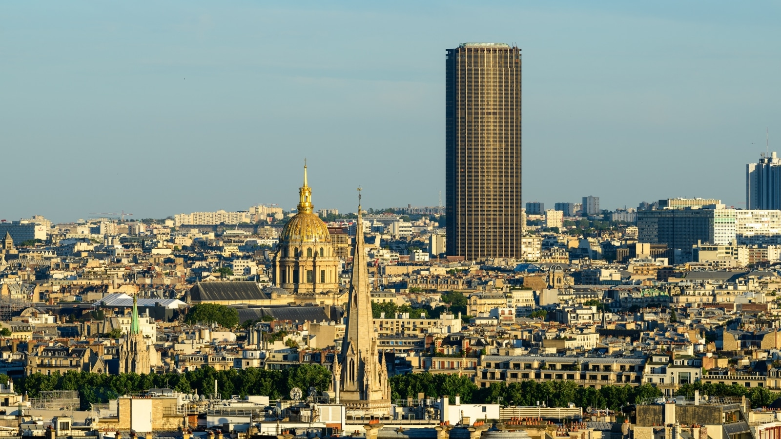 The image depicts Paris cityscape featuring La Tour Montparnasse, a prominent building with an observation deck. The structure stands out amidst the urban landscape, showcasing modern architectural design. Surrounding the tower are other buildings and city structures, reflecting Paris's mix of historic and modern elements.