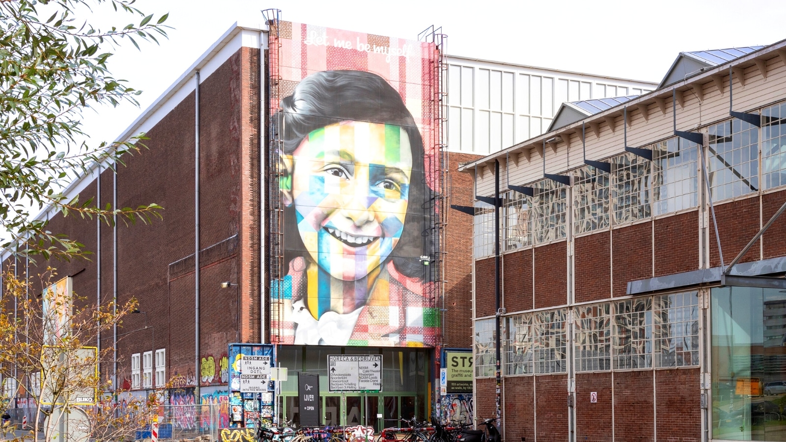 The image captures a street art scene in Amsterdam, Netherlands, focusing on the famous mural titled "Let Me Be Myself," depicting Anne Frank. The mural is prominently displayed on a wall, showcasing artistic elements and cultural significance.