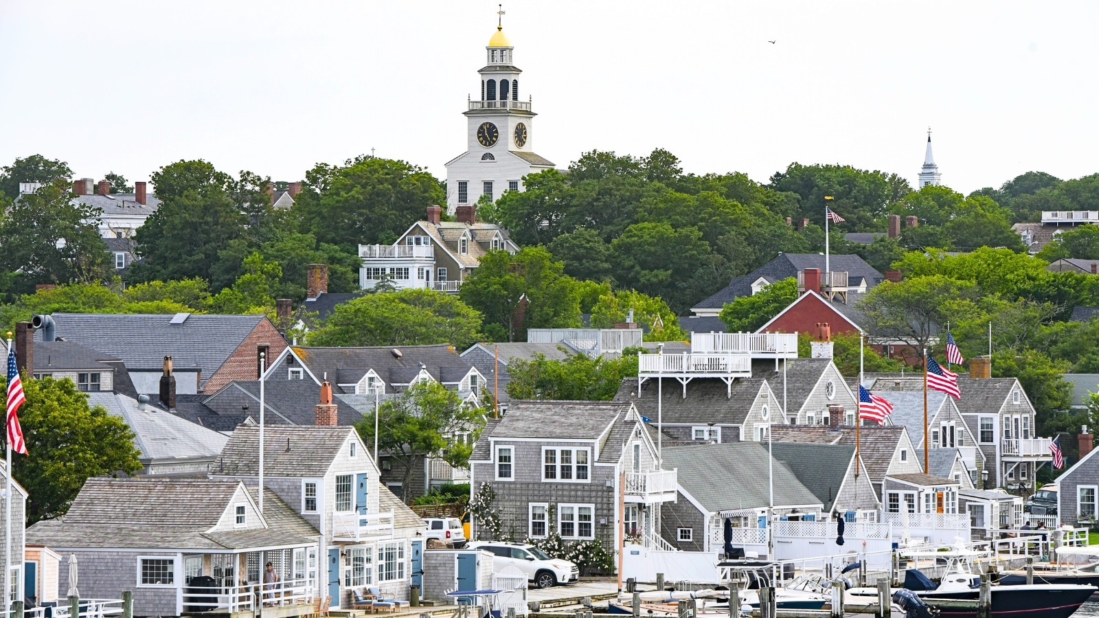 The image captures a view of Nantucket Town from the ferry in Nantucket Harbor. The scene includes several yachts docked at the pier. The houses are closely situated, typical of traditional coastal architecture, with a church visible at the higher elevation of the town.