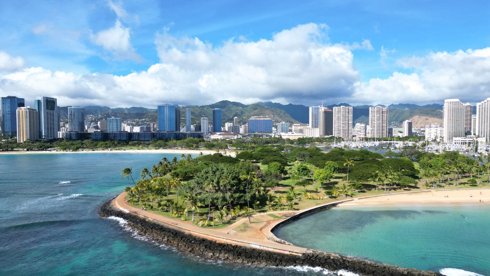 The image captures the coastline of Magic Island in O'ahu, Hawaii. In the foreground, there is a small island featuring palm trees and a pathway for visitors. In the background, a larger island is visible, characterized by high-rise buildings that contrast with the more natural surroundings of the smaller island.